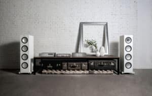 White KEF loudspeakers standing at either end of a coffee table with a white brick wall behind it.