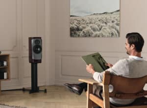 A man is listening to a vinyl album while seated on a chair, through a Dynaudio standmount speaker placed in the front of the room.