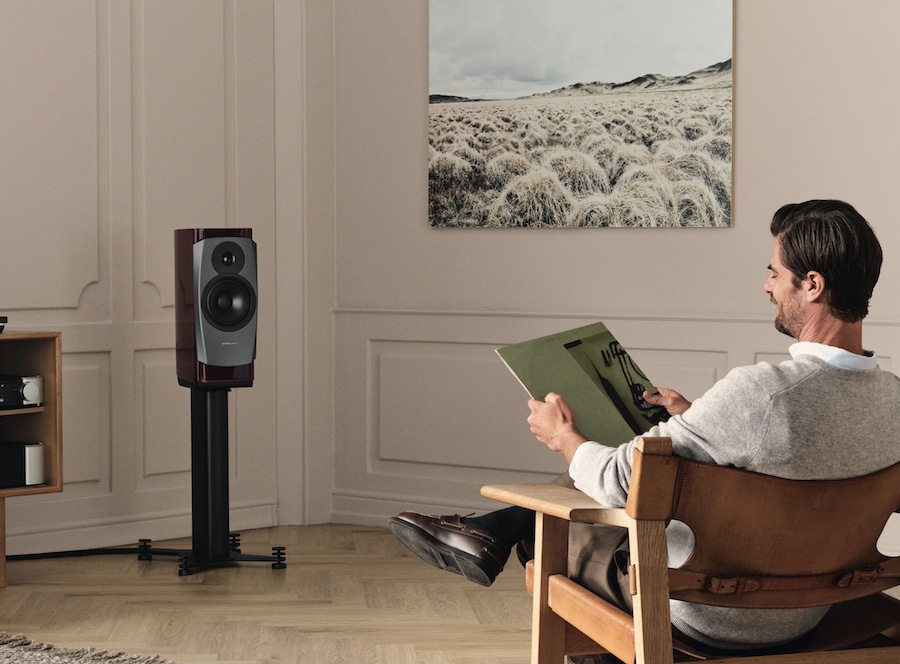 A man is listening to a vinyl album while seated on a chair, through a Dynaudio standmount speaker placed in the front of the room.
