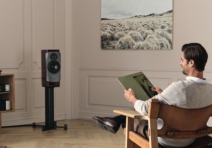 A man is listening to a vinyl album while seated on a chair, through a Dynaudio standmount speaker placed in the front of the room.