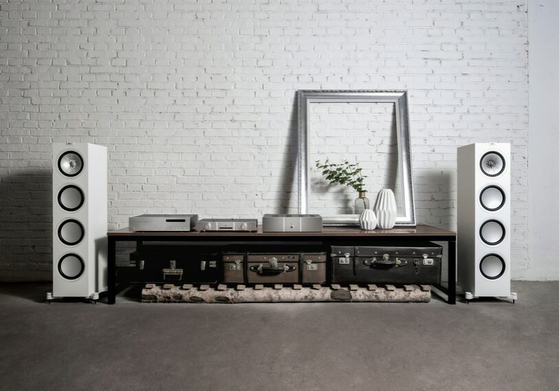 White KEF loudspeakers standing at either end of a coffee table with a white brick wall behind it.